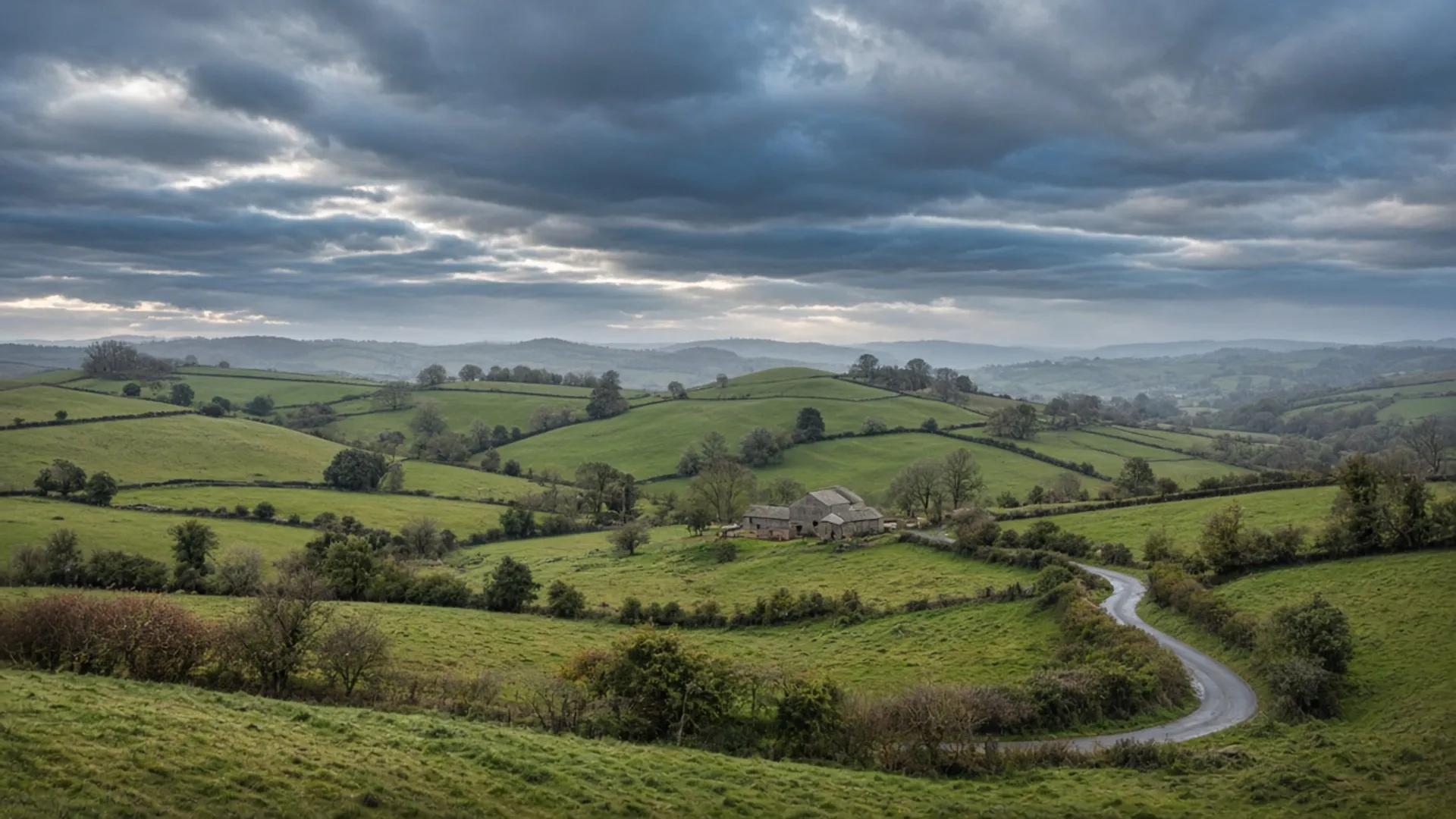 Rolling British fields, hedgerows and a distant stone farmstead under broken slate-blue cloud